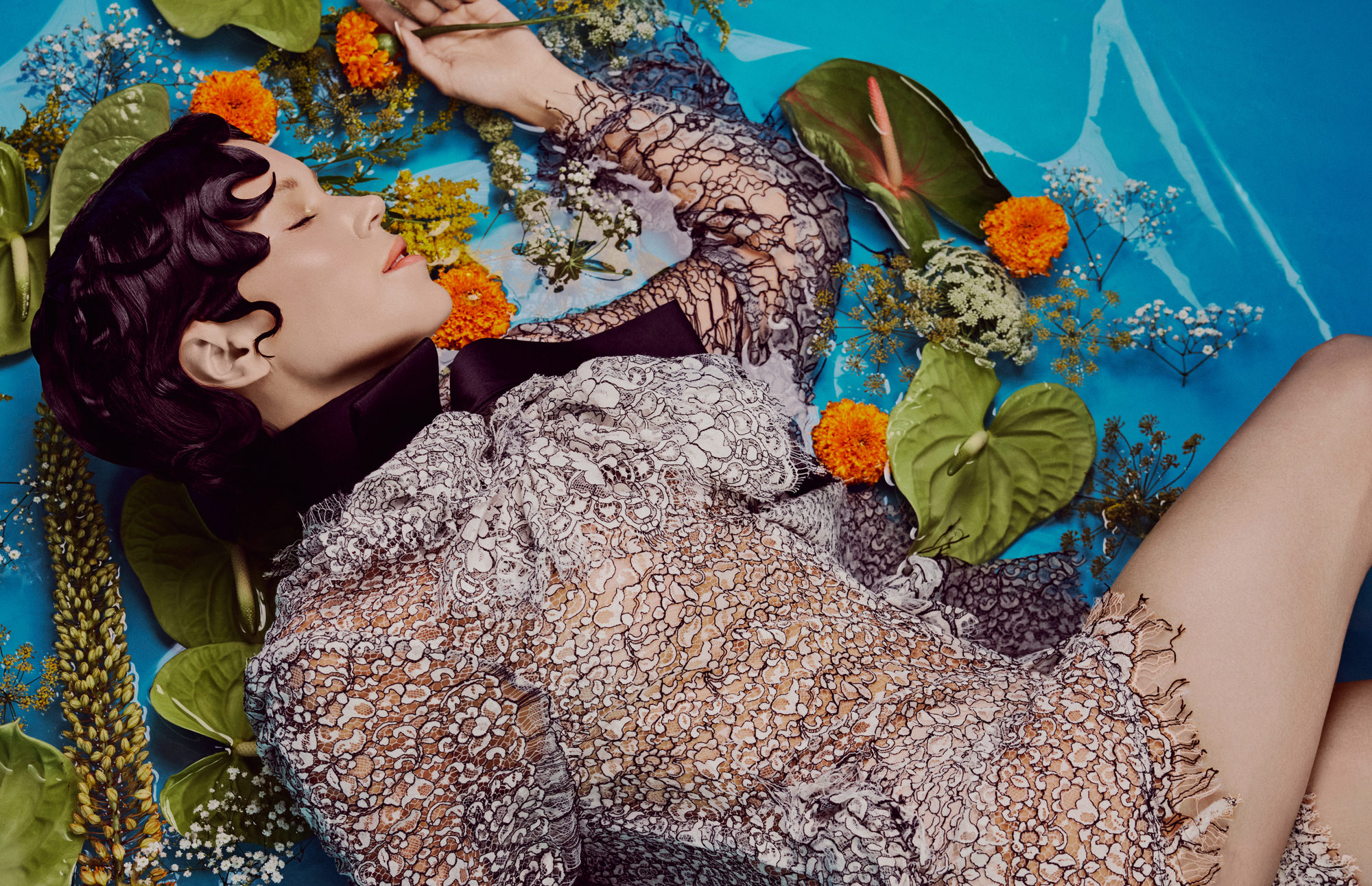 A woman wearing an intricate lace dress floats in vibrant blue water surrounded by colorful flowers and foliage.