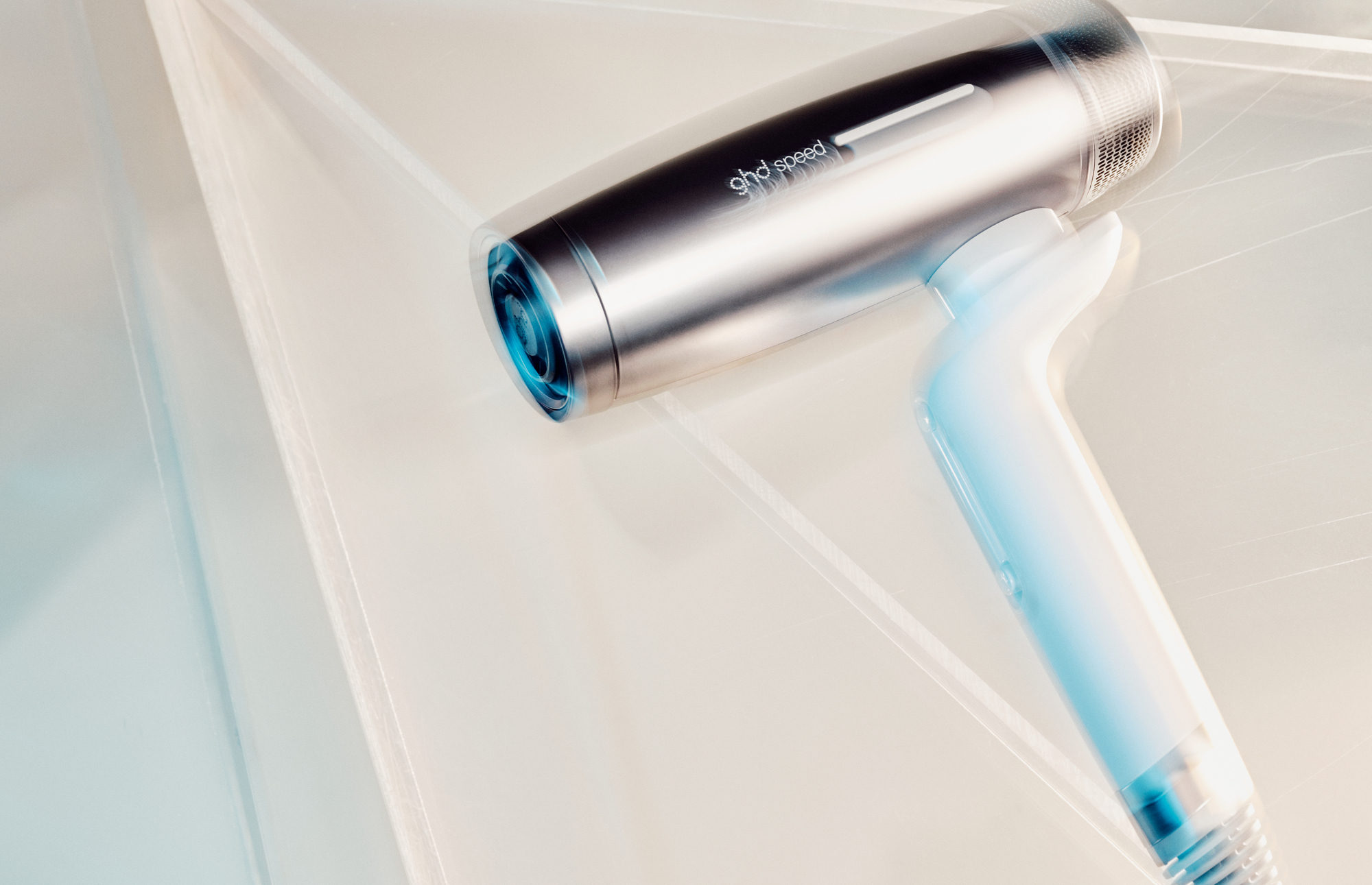 A modern ghd speed hair dryer with a metallic barrel and translucent handle resting on a minimalist geometric surface.