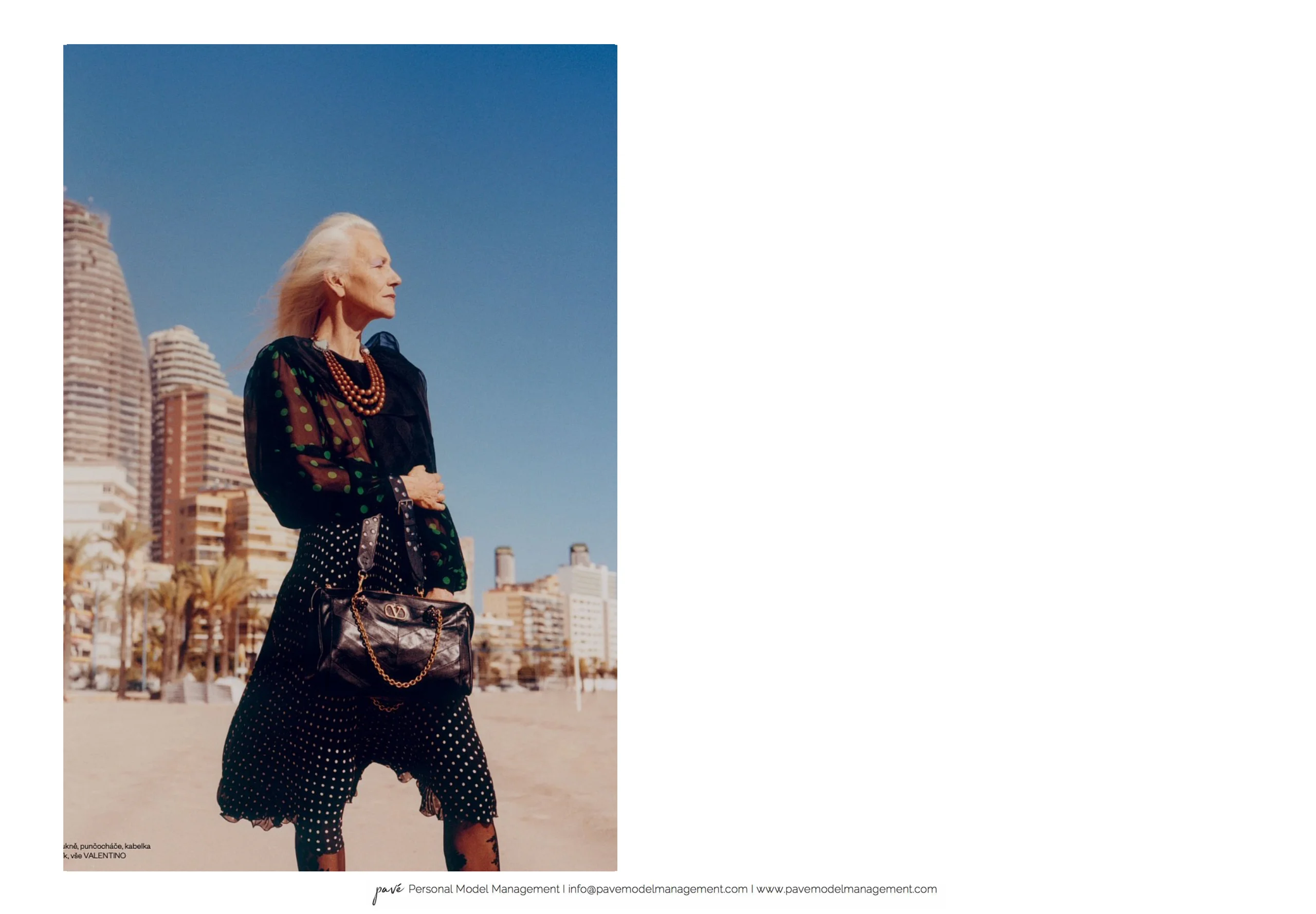 An older woman with blonde hair and a beaded necklace stands on a sandy beach, holding a purse and looking toward city buildings.