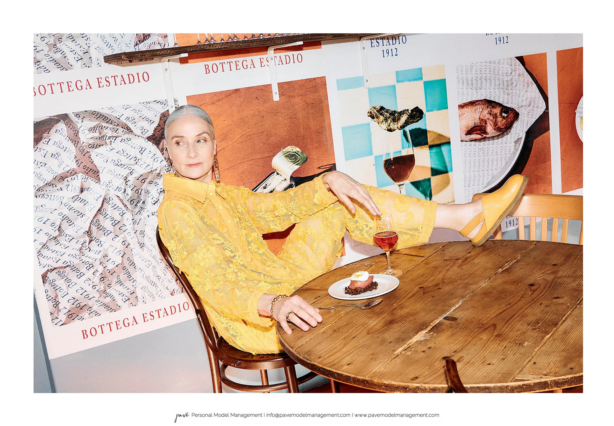 A model reclines at a table enjoying wine and dessert, dressed in a yellow lace outfit against a backdrop of Bottage Estadio posters.