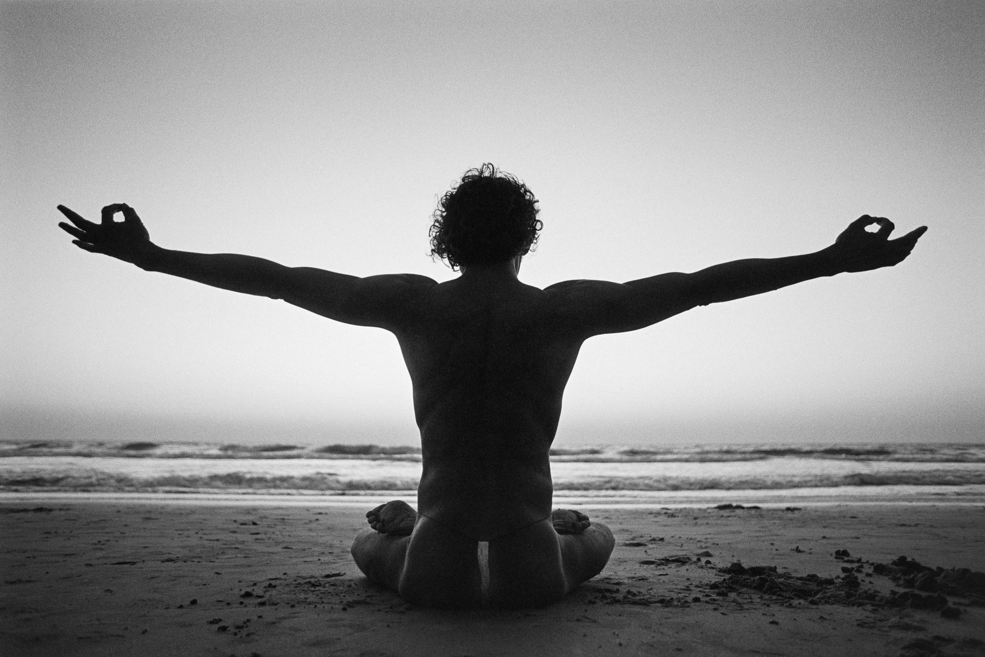 Black-and-white photograph of a man sitting on a beach facing the sea, seen from behind with his arms extended.